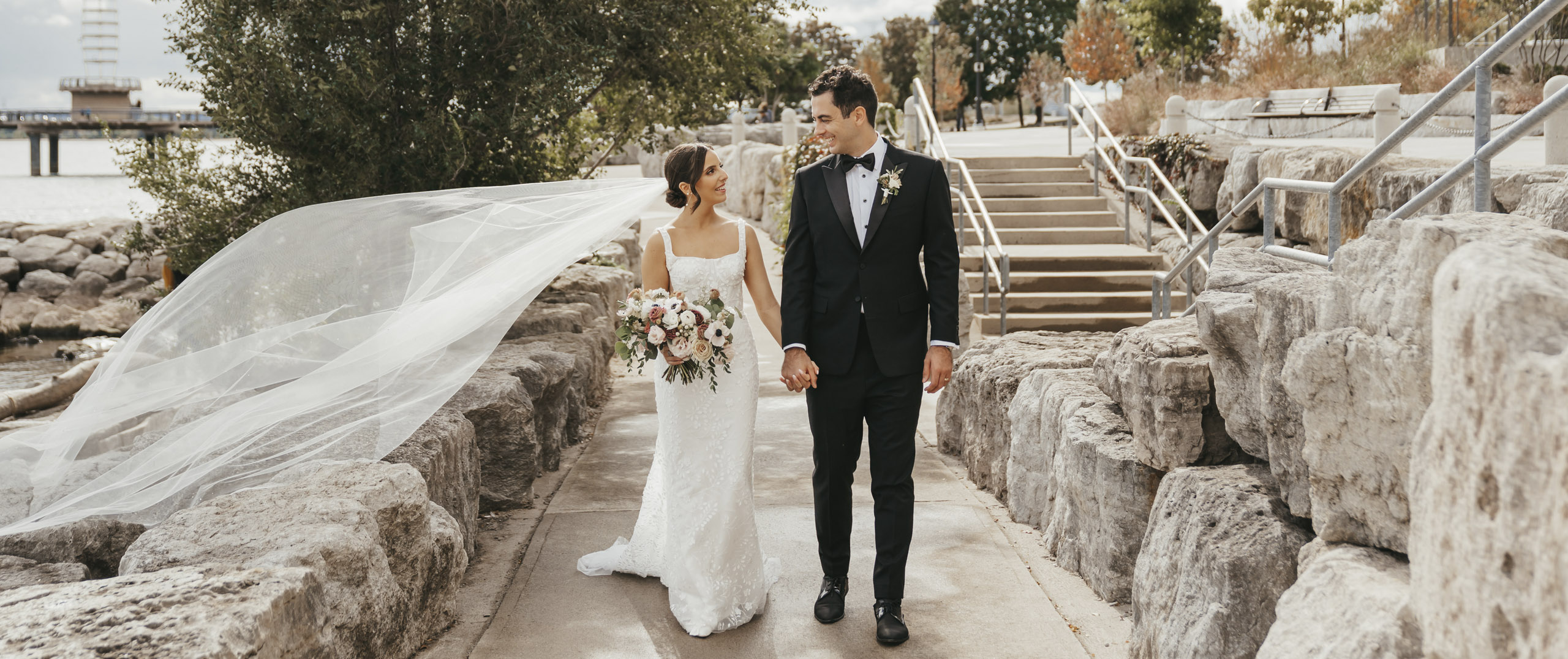 Couple walking alongside lake side side walk, with her bridal train blowing in the wind